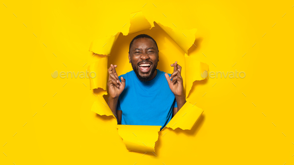 Wishing good luck. Emotional black man gesturing and crossing fingers, posing through hole in torn