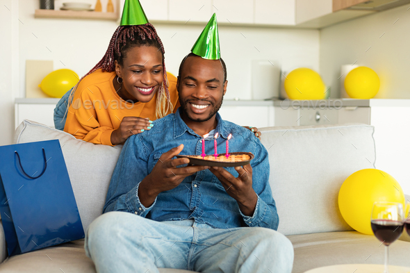 Happy african american wife surprising her husband with handmade birthday pie, man blowing candles