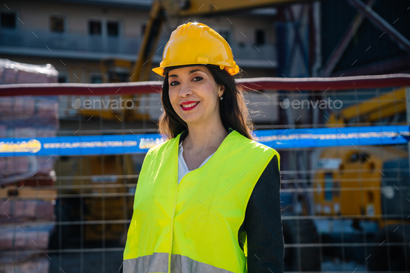 Smiling ethnic female worker in hardhat standing near building during construction
