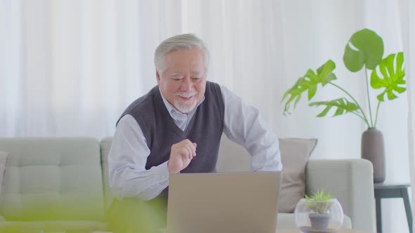 elderly asian man sitting on sofa and using computer laptop VDO Conference online meeting alt