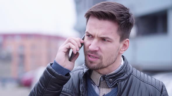 A Young Handsome Man Argues with Someone on a Smartphone in a Suburban Area alt