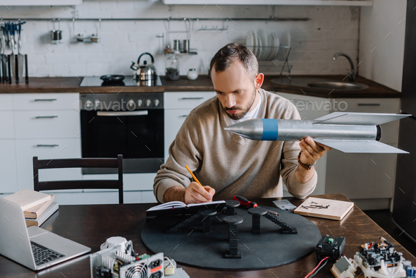 handsome engineer holding rocket model and making notes at home