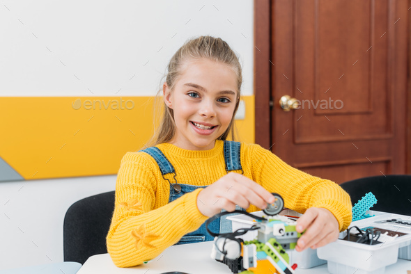 cheerful schoolgirl looking at camera and working on handmade robot model during STEM lesson