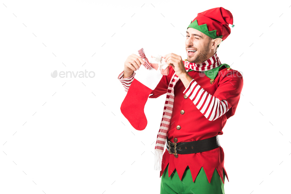man in christmas elf costume winging and putting present in red christmas stocking isolated on white