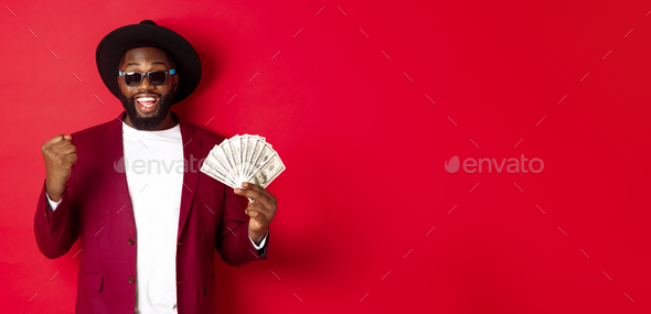 Cheerful african american guy in sunglasses, black hat and blazer, winning prize money, holding