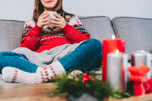 Cropped woman in ugly sweater and warm woolen socks on sofa holding cup of hot drink m