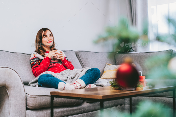Smiling Woman in xmas ugly sweater and warm woolen socks on sofa holding cup of hot drink