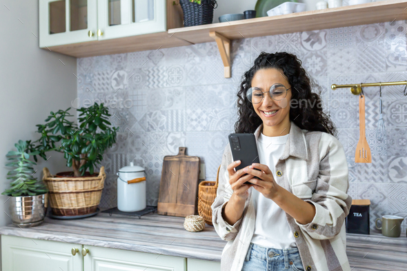 Young hispanic woman standing at home in kitchen with phone. Makes online purchases, orders