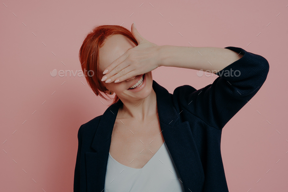 Cute positive ginger woman holding hand in front of face, covering eyes in anticipation for surprise