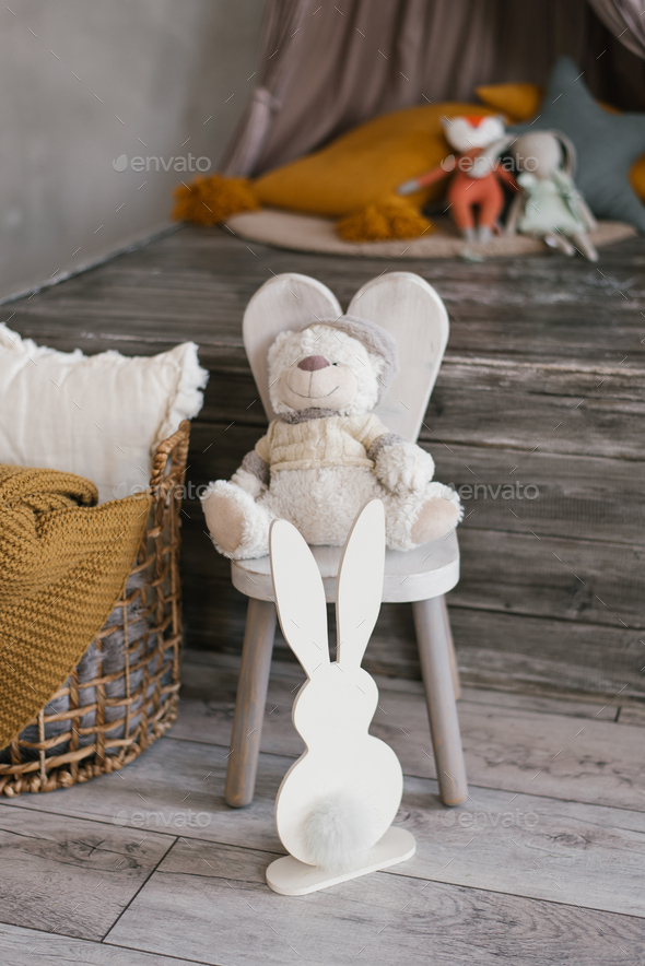 Children\'s soft toy bear sits on a chair, next to it there is a plywood bunny and a basket