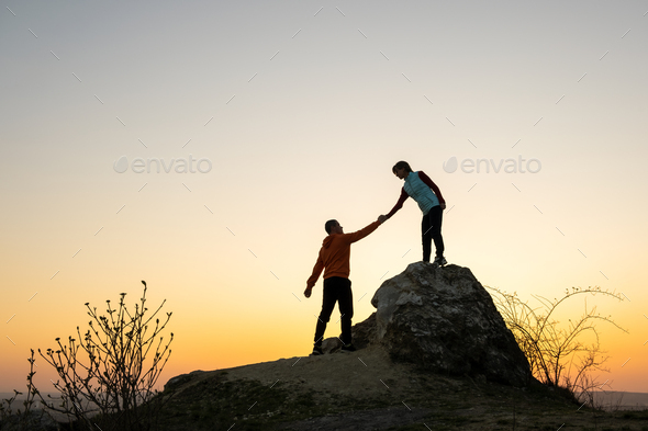 Man and woman hikers helping each other to climb a big stone at sunset in mountains.