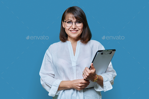 Middle aged business woman with clipboard looking at camera on blue background - Stock Photo - Images