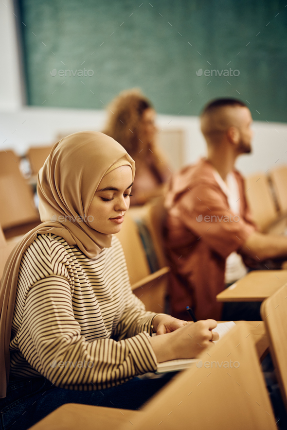 Muslim female student taking notes while attending a class at college classroom.