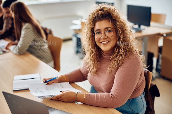 Happy female student having an exam at college classroom and looking at camera.