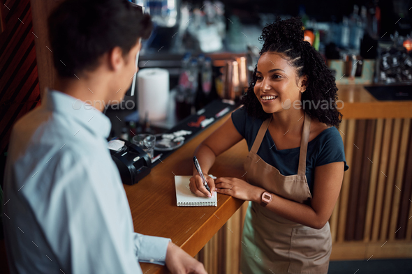 Happy black waitress communicating with coworker while taking notes at bar counter.