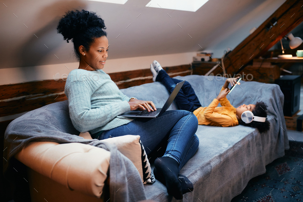 Happy black woman using laptop while daughter using using smart phone at home. - Stock Photo - Images