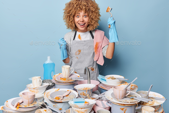 Happy busy woman with curly hair wears apron does dishwashing stands near sink full of dirty dishes