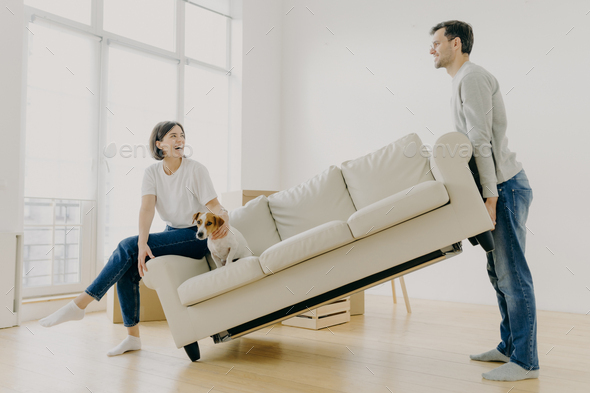 Indoor shot of funny family couple furnish their first home, carry big white sofa - Stock Photo - Images