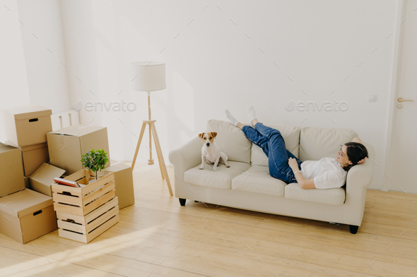 Female renter lies on white comfortable couch, raises crossed legs, poses with favourite dog - Stock Photo - Images
