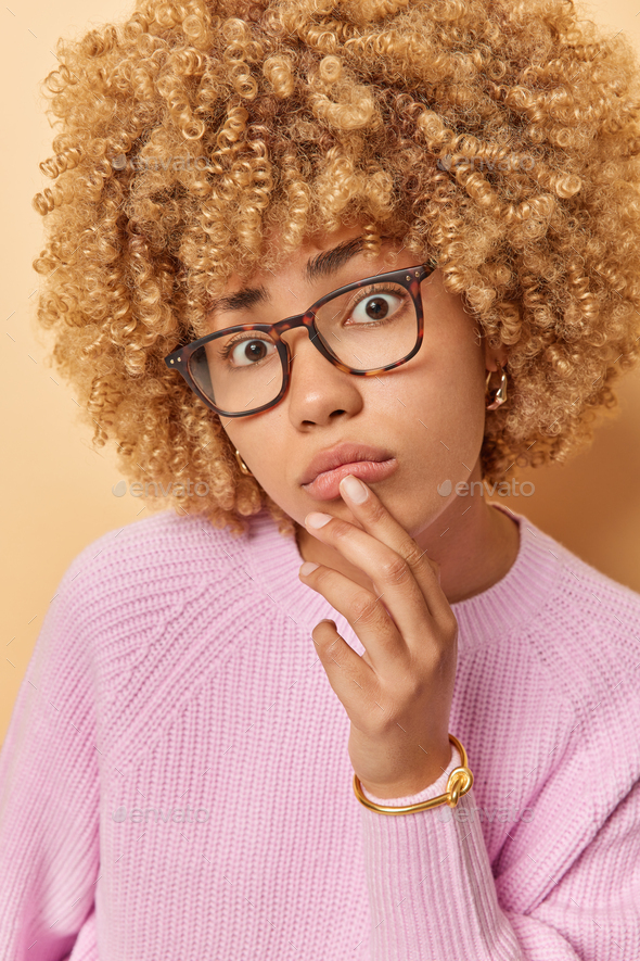 Close up shot of frustrated sad curly woman looks with wonder at something touches lips wears - Stock Photo - Images