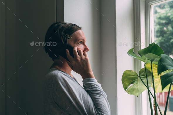 Sad alone woman talking on mobile phone while looking out the window of living room - Stock Photo - Images