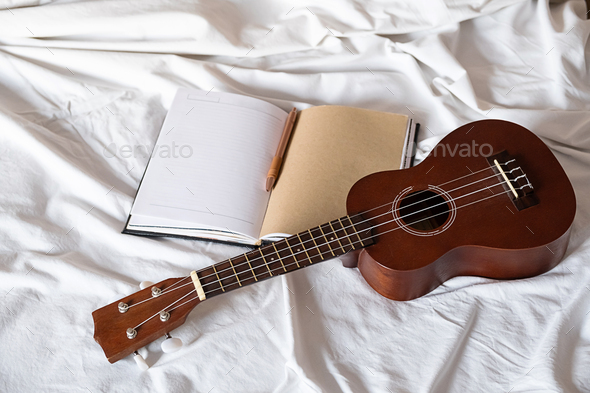 Ukulele guitar and open notebook on white bed. Cozy atmosphere for writing music.