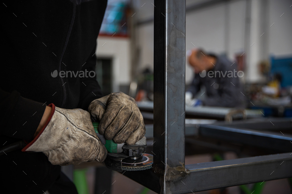 Close-up of the hands of a worker using the sander on a metal bar while wearing protective gloves.