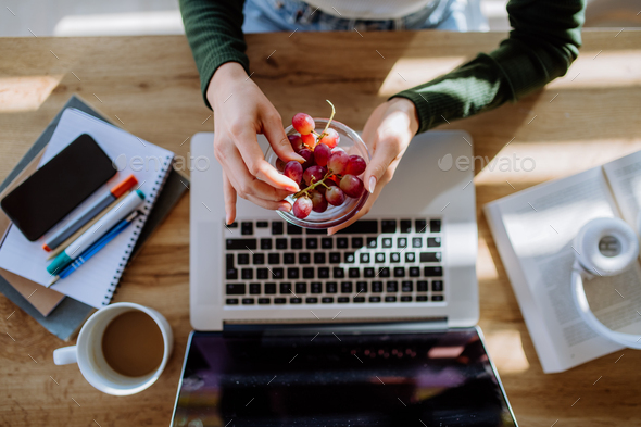 Top view of woman holding bowl with grapes above desk with computer, diary and smartphone. Work-life - Stock Photo - Images