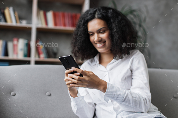 Attractive young woman in casual wear sitting on comfy couch with modern smartphone