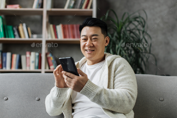 Attractive asian man in casual wear sitting on comfy couch with modern smartphone in hands.