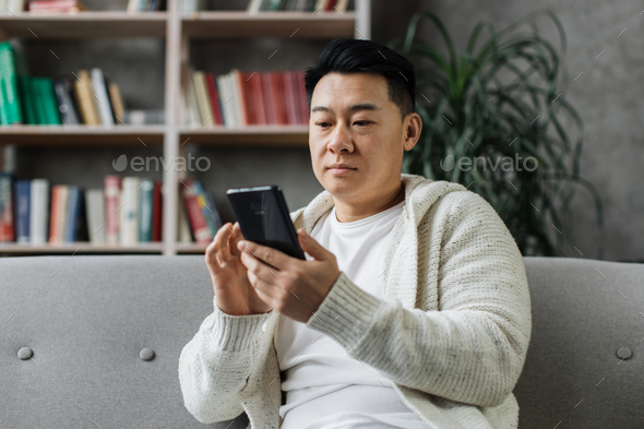 Attractive asian man in casual wear sitting on comfy couch with modern smartphone in hands.