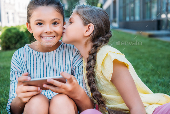 adorable little schoolgirl with smartphone looking at camera while her friend kissing her cheek