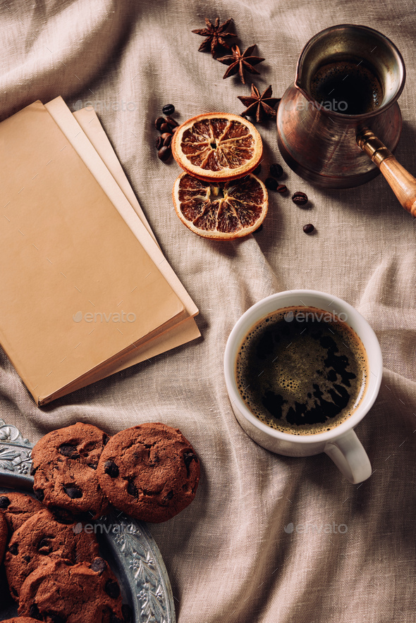 top view of cup of coffee with book and chocolate chip cookies on beige cloth