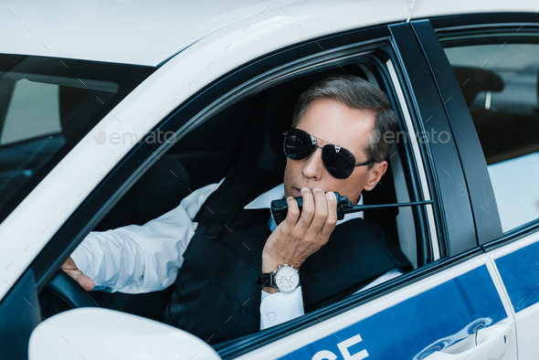 middle aged male police officer in sunglasses talking on radio set in car