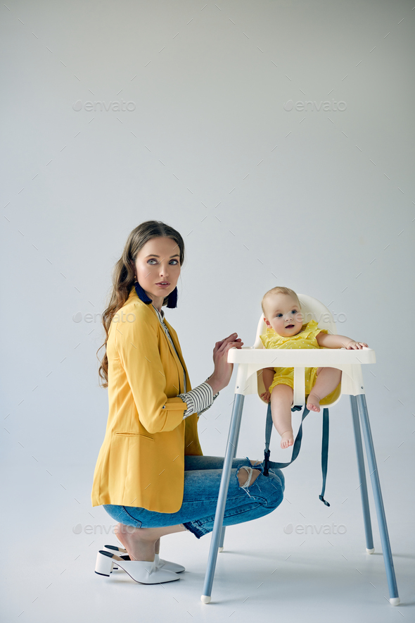 beautiful fashionable mother crouching near infant daughter sitting in high chair on grey