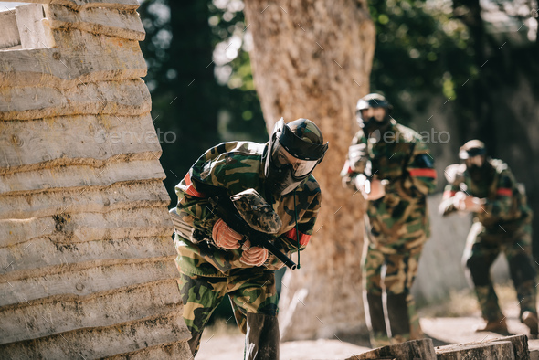 selective focus of paintball player in protective mask holding marker gun looking out of wooden wall