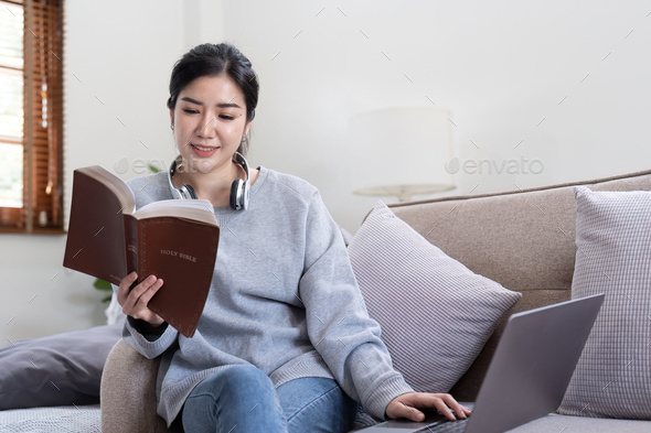 Young beautiful asian woman student sitting on sofa reading book at home while using laptop for - Stock Photo - Images