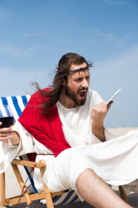 angry Jesus sitting on sun lounger with glass of wine and screaming at smartphone in desert