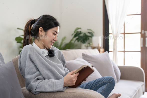 Young beautiful asian woman student sitting on sofa reading book at home. - Stock Photo - Images