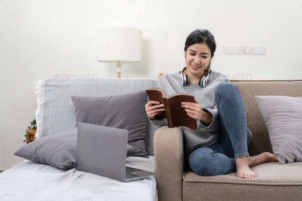 Young beautiful asian woman student sitting on sofa reading book at home while using laptop for - Stock Photo - Images