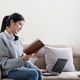 Young beautiful asian woman student sitting on sofa reading book at home while using laptop for - PhotoDune Item for Sale