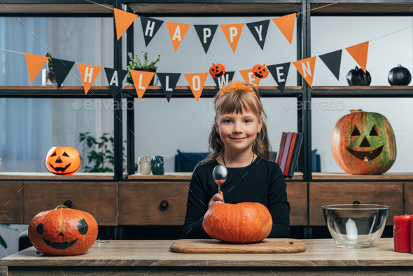 portrait of smiling kid at tabletop with pumpkins and hanging happy halloween flags behind at home - Stock Photo - Images