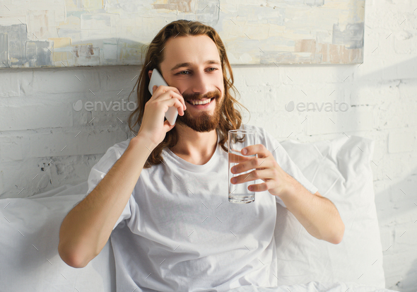 cheerful bearded man with long hair talking on smartphone and drinking water in bed during morning - Stock Photo - Images