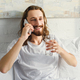 cheerful bearded man with long hair talking on smartphone and drinking water in bed during morning - PhotoDune Item for Sale