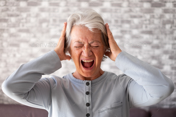 Desperate frustrated senior woman screaming in pain with hands on ears, standing at home alone