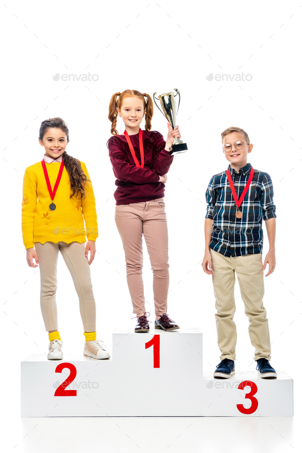 smiling preteen kids with medals and trophy cup standing on winner pedestal, smiling and looking at