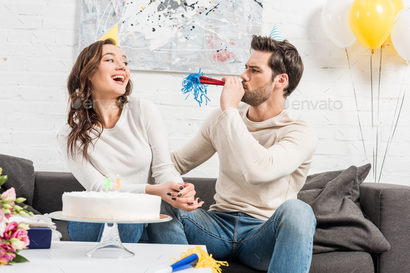 couple in party hats celebrating birthday with cake while man blowing party horn at home