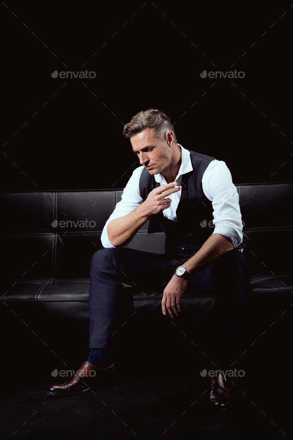 serious handsome man in formal wear sitting on couch with cigarette isolated on black