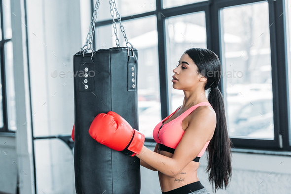 side view of young african american sportswoman in boxing gloves holding punching bag in gym