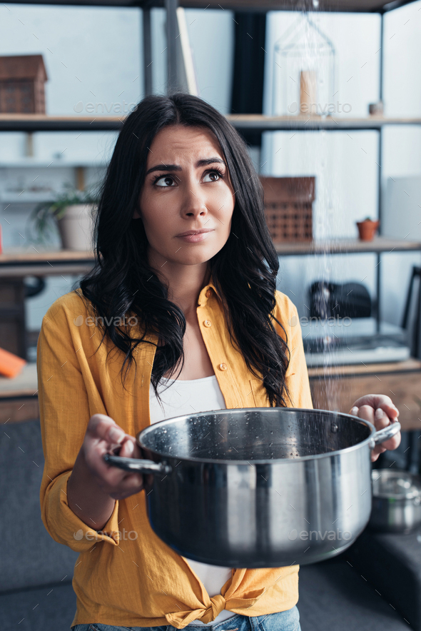 Sad girl in yellow shirt holding pot under water leaking from ceiling